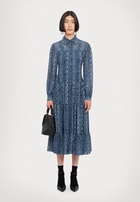 Rochie albastră cu mâneci lungi, imprimeu cu șarpe, guler și fustă în straturi. Asortată cu o geantă neagră și ciorapi negri până la glezne, cu tocuri.