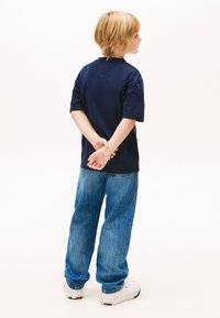 Tricou bleumarin cu mâneci scurte, lejer, asortat cu blugi albastru deschis. Adidași albi cu accente roșii și albastre. Copilul stă cu mâinile la spate.