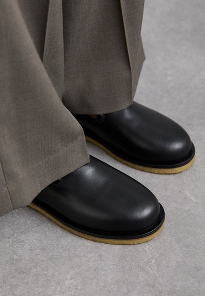 Scarpe in pelle nere con punta arrotondata, suola piatta e suola esterna di un marrone chiaro a contrasto, abbinate a pantaloni grigi larghi.
