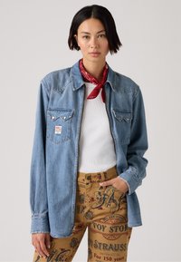 Lys blå denimskjorte med spiss ståle, brystlommer og sølvknapper. Kombinert med brune bukser med røde grafiske trykk. Rød bandana som tilbehør.