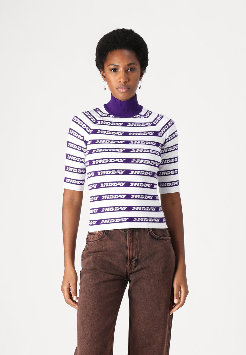 Maglione a collo alto bianco e viola a righe con testo "2NDAY", maniche corte, abbinato a jeans scuri a vita alta. Texture liscia.