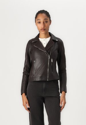Femme aux cheveux tressés portant une veste en cuir marron foncé zippée de manière inégale sur une chemise blanche boutonnée et un pantalon noir.