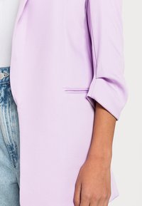 Blazer lavande avec une texture lisse, un tissu doux et des manches retroussées. Comprend un petit détail de poche latérale pour un intérêt esthétique.