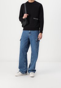 Persona vistiendo un sudadera negra de Calvin Klein, jeans cargo azules, zapatillas blancas y un bolso cruzado negro contra un fondo blanco liso.