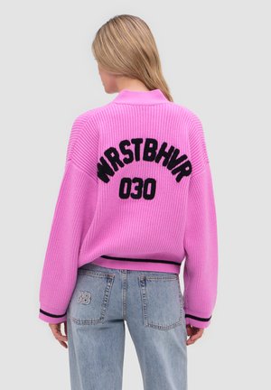 Kobieta z długimi blond włosami, ubrana w jaskraworóżowy sweter z napisem „WRSTBHVR 030”, do tego jasnoniebieskie dżinsy z naszywką „WB”.