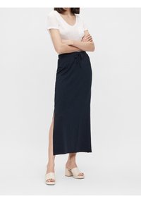 Femme debout les bras croisés, portant un haut blanc à manches courtes, une jupe longue bleu marine avec une fente latérale et des sandales beiges à talon bloc et à bout ouvert.