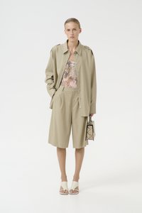 Chemise beige oversize à boutons, haut à motifs, shorts ajustés, sandales blanches à bout ouvert et un petit sac à main en serpent.