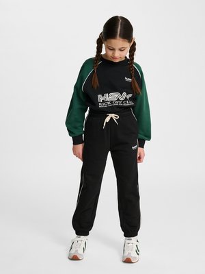 Ung pige med flettet hår iført sort og grøn sweatshirt og sorte joggingbukser med hvide sneakers, kigger ned.