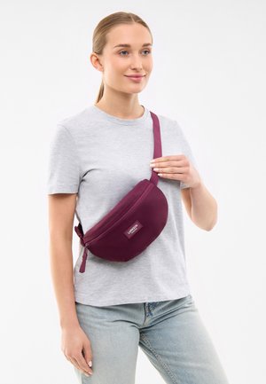 Donna con una maglietta grigia chiaro e jeans blu chiaro, con una borsa a tracolla viola tenuta dal suo manico diagonale sul petto.