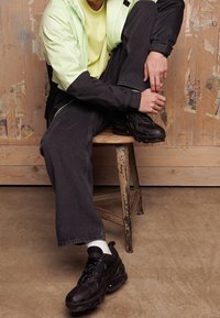 Černé sportovní tenisky s texturovanou podrážkou, nošené s tmavými džínovými kalhotami, světle žlutou košilí a mátově zelenou bundou, sedící na stoličce.