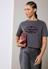 Graues kurzärmliges Crop-T-Shirt mit dem Text "LA PARISIENNE EST. 1982" in dunklem Weinrot, kombiniert mit einem glänzenden schwarzen Rock und einer weinroten Clutch.