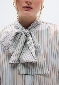 Ριγέ μπλούζα σε ανοιχτές αποχρώσεις με μπλε και καφέ γραμμές· διαθέτει μεγάλο φιόγκο στον λαιμό. Ομαλή υφή, άνετη εφαρμογή.