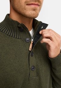 Strikket olivengrøn sweater med ribbet krave, lynlås lukning og to knapper. Har et struktureret mønster og læderdetalje på lynlåsens træk.  