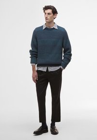 Πλεγμένο σκούρο τιρκουάζ πουλόβερ με ριγωτή υφή, φορεμένο πάνω από ένα λευκό και μαύρο καρό πουκάμισο. Συνδυασμένο με μαύρο παντελόνι και δερμάτινα παπούτσια.