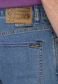 Jeans de denim azul con un parche de cuero marrón texturizado que presenta un diseño en relieve y un pequeño logo negro de "Volcom" en el bolsillo trasero.
