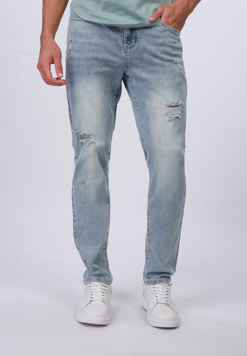 Jeans de mezclilla de color azul claro con acabados desgastados, que presentan rotos visibles, corte recto, cinco bolsillos y un leve desteñido, acompañados de zapatillas blancas.