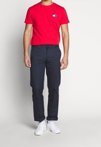 Czerwona bawełniana koszulka z małym logo na piersi, połączona z granatowymi spodniami i białymi sneakersami. Gładka tekstura, standardowy okrągły dekolt, luźny krój.