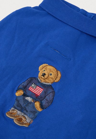 Polo azul con un oso de peluche bordado que lleva un atuendo de mezclilla y un suéter con la bandera estadounidense en el pecho.