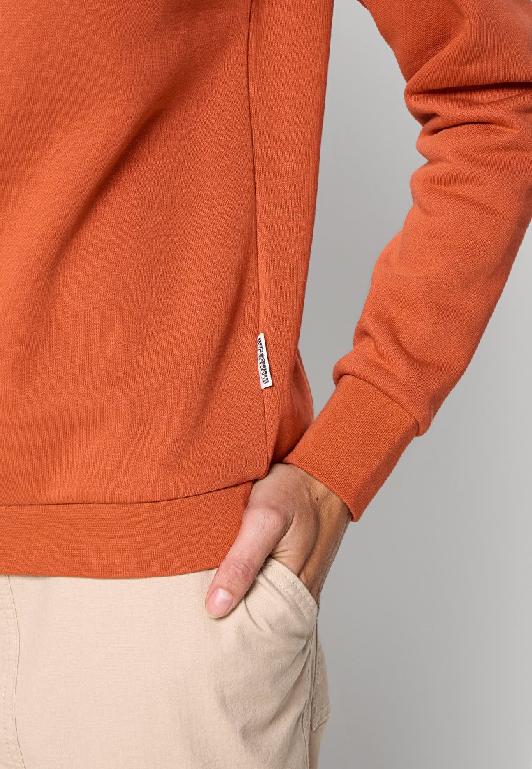 Osoba ubrana w pomarańczową bluzkę z długim rękawem z małą białą metką z boku, ręka swobodnie włożona w kieszeń beżowych spodni.
