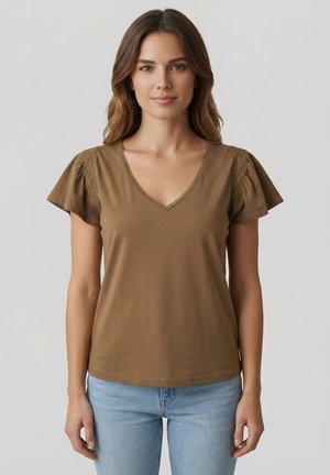 Jeune femme aux longs cheveux bruns portant un chemisier marron à col en V avec des détails en dentelle sur les manches courtes et un jean bleu clair, debout devant un fond gris.