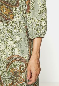 Zelené šaty s vzorem, které obsahují složité květinové a paisley designy. Měkký materiál s volným střihem a nařasenými balónovými rukávy.