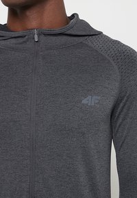 Szara bluza z kapturem zapinana na zamek, z teksturowanym wzorem na ramionach, z logo na piersi i gładkim wykończeniem tkaniny.