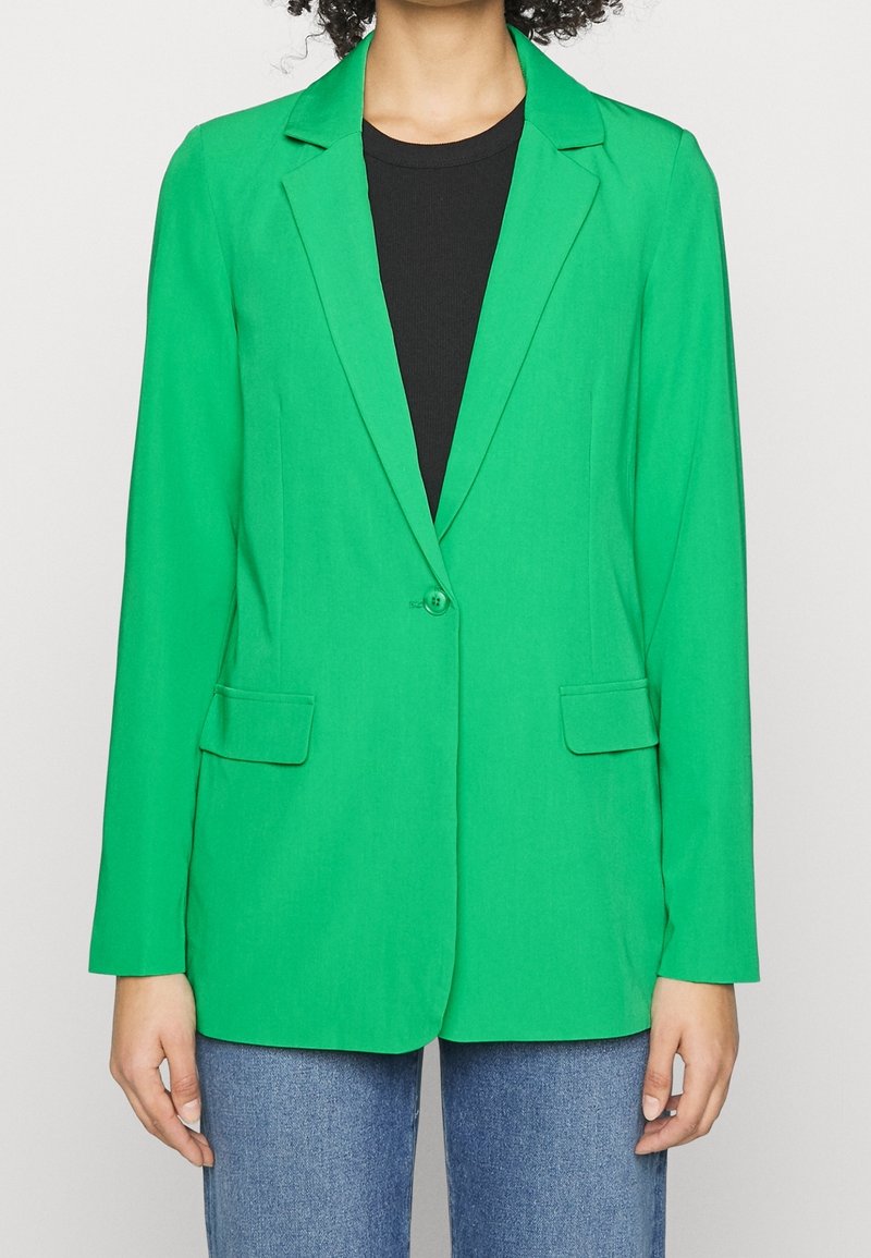 Personne portant un blazer vert vif à un seul bouton sur un top noir et un jean bleu clair, raccourci au niveau du cou et au milieu de la cuisse.