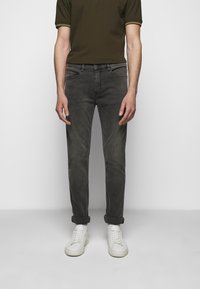 Mężczyzna w podwijanych ciemnoszarych dżinsach, białych sneakersach, białych skarpetkach i oliwkowej koszulce polo z krótkim rękawem, stojący przy białej ścianie.