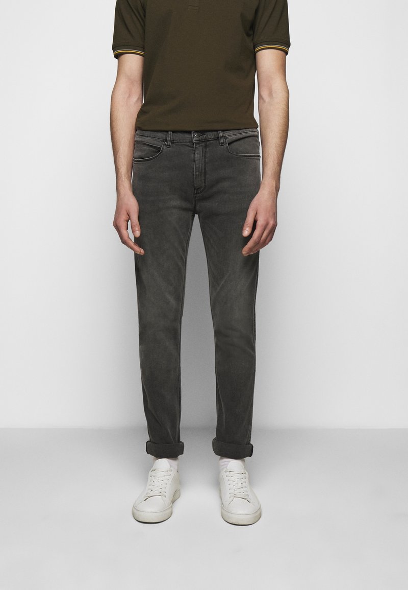 Mężczyzna w podwijanych ciemnoszarych dżinsach, białych sneakersach, białych skarpetkach i oliwkowej koszulce polo z krótkim rękawem, stojący przy białej ścianie.