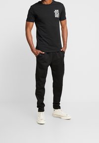 Camiseta de algodón negra con texto blanco en el pecho, combinada con joggers negros. Los joggers tienen bolsillos y un diseño ajustado. Zapatillas blancas.