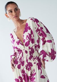 Õrnas toonis kreemikas lilleline kleit, kaunistatud suurte lillaõitega, sügava V-kaelusega ja kokku kogutud vöökohaosaga. Pikkade põrkavatele varrukad lisavad mahtu ja tekstuuri.