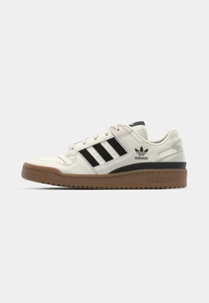 Zapatilla de cuero blanca con rayas negras, suela de goma y cuello acolchado. Presenta el logo de Adidas en la lengüeta y en el lateral. Diseño clásico de corte bajo.