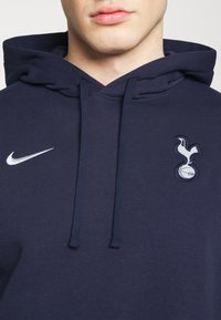 Gros plan d'une personne portant un sweat à capuche bleu marine avec le logo Nike à gauche et l'emblème du club de football Tottenham Hotspur à droite.