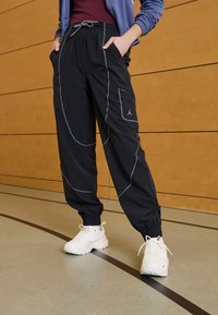 Pantalones de chándal negros con costuras blancas, que presentan una cinturilla elástica y bolsillos laterales. Combinados con zapatillas blancas, con un fondo de madera.