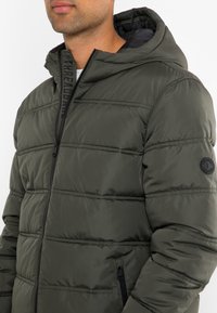 Zielona oliwkowa kurtka puchowa z zamkiem błyskawicznym z przodu, poziomymi panelami pikowanymi, czarnymi dodatkami oraz łatą z logo na lewym rękawie. Model z kapturem.