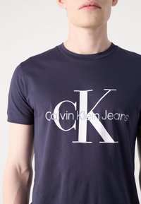 Férfi sötétkék pólóban, elöl nagy fehér "cK" betűkkel és kisebb "Calvin Klein Jeans" szöveggel.