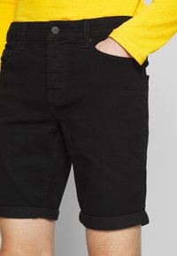 Shorts de mezclilla negros con dobladillo enrollado, diseño de cinco bolsillos, cierre de botón metálico y una textura suave. Llevados con una camiseta de manga larga amarilla.