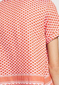 Oranžovo-bíle vzorovaná tkanina s geometrickými a pruhovanými vzory pokrývá část horního ramene a ramene osoby.