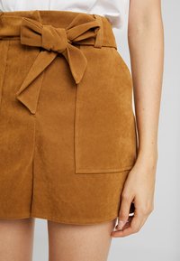 Brun mockaskjol med en framsidan knytband och två sidofickor, med en mjuk textur och ren sömnad. Sömmen sitter ovanför låret.