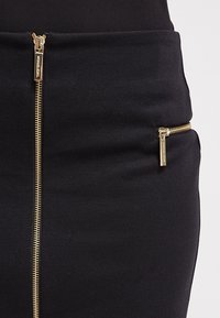 Jupe noire en tissu lisse avec des zips dorés à l'avant et sur le côté pour des détails fonctionnels.
