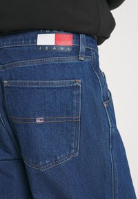 Mørkeblå denimjeans med oransje sømmer, med et merkelapp i rødt, hvitt og blått på linningen og en liten logo på baklommen.