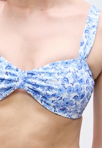 Κοντινό πλάνο ενός ατόμου που φοράει μπλε και άσπρο μπικίνι με floral μοτίβο, φαρδιές τιράντες στους ώμους και κόμπο στη μέση.