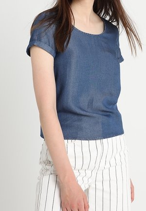 Žena oblečená v modrém tričku s krátkými rukávy a bílých kalhotách s černými vertikálními pruhy stojící před jednobarevným pozadím.