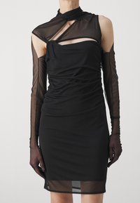 Svart klänning med figursydd siluett, med genomskinliga långa ärmar och utskärningar. Strukturerad tyg med samlade detaljer.