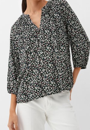 Kvinde iført sort blomsterbluse med indsamlet halsudskæring og trekvartærmer, kombineret med hvide bukser.