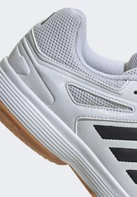 Zapatilla deportiva blanca con parte superior de malla, acentos de rayas negras, suela de goma texturizada y refuerzo en el talón. Presenta un diseño suave y transpirable.
