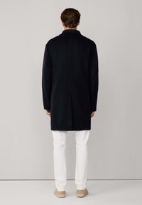 Uomo con un lungo cappotto nero, pantaloni bianchi e scarpe beige, in piedi di fronte a una parete grigia chiaro uniforme.