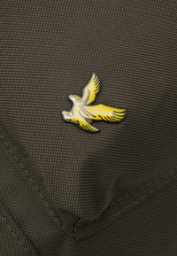 Spilla a forma di uccello giallo su uno sfondo di tessuto verde scuro e testurizzato. La spilla presenta un design dettagliato con le ali spiegate e contorni neri.