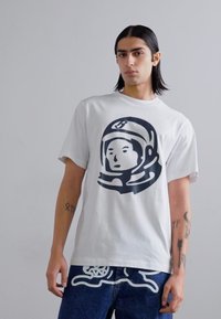 White cotton t-shirt featuring a large black astronaut graphic on the front, short sleeves, and a relaxed fit. Denim jeans with white accents.