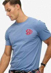Hombre vistiendo una camiseta de manga corta de color azul claro con un logo rojo "DCo" en el pecho, una camisa de cuadros atada alrededor de la cintura y un reloj de pulsera negro.